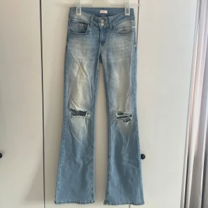 Ljusblå bootcut jeans - Säljer ett par ljusblå bootcut jeans från Nelly med slitna detaljer på båda knäna och snygg tvätt. Jeansen har fem fickor, dubbla knappar i midjan och bakfickor med lock och knapp. Perfekta för en avslappnad och trendig look. Aldrig använda, endast testade på. Innerbenslängd: 84
