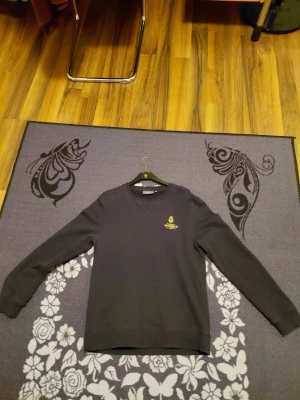 Svart sweatshirt från Morris - Snygg Mörkblå/Svart sweatshirt från Morris med gul broderad logga och text på bröstet. Klassisk rund halsringning och långa ärmar. Perfekt för dig som gillar stilrena och tidlösa plagg med en sportig touch.