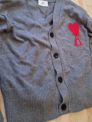 Grå kofta från Ami Paris med rött hjärta - Grå kofta från Ami Paris med v-ringning och stora svarta knappar. På bröstet finns ett rött hjärta och bokstaven A. Koftan har bred ribbstickad kant nedtill och vid ärmsluten. Perfekt för dig som gillar stilrena plagg med en twist.