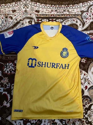 Al Nassr Ronaldo matchtröja gul/blå - Säljer en gul och blå Al Nassr matchtröja med Ronaldo och nummer 7 på ryggen. Tröjan har klubbmärke och sponsortryck på bröstet samt tryck på båda ärmarna. Tillverkad i lätt och ventilerande material, perfekt för fotbollsträning eller match.