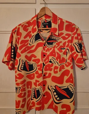 Santa Cruz/Urban Outfitters skjorta - Mönstrad skjorta från Santa Cruz/Urban Outfitters, med korta ärmar i orangerött, beige och mörkblått. Skjortan har klassisk krage, bröstficka och knäppning framtill.