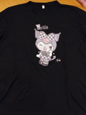 Gullig Svart oversized t-shirt med Kuromi-tryck - Säljer en super cutsie oversized svart t-shirt med ett stort tryck av Kuromi på framsidan. Motivet har rutiga detaljer i lila och vitt samt texten 'cutie' ovanför. Perfekt för dig som gillar kawaii-stil och vill sticka ut lite extra. Mycket använd och älskad av mig i 6:an men är endå i bra skick. Har inte använt sen dess. Priset kan diskuteras
