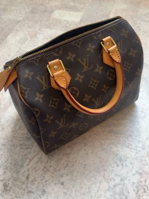 Väska - Klassisk Louis Vuitton Speedy handväska i brunt monogrammönster med beige läderhandtag och guldfärgade metalldetaljer. Väskan har dragkedja upptill och ikoniska LV-loggor över hela ytan. Perfekt för dig som vill ha en tidlös och lyxig accessoar.