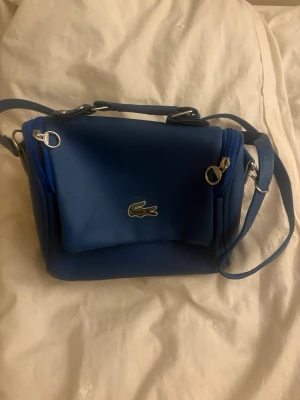 Blå axelremsväska från Lacoste - Snygg blå handväska från Lacoste med justerbar axelrem och silverfärgade detaljer. Väskan har ett lock med magnetknäppning och det klassiska krokodilmärket framtill. Tillverkad i slätt konstläder och har flera fack för smart förvaring.