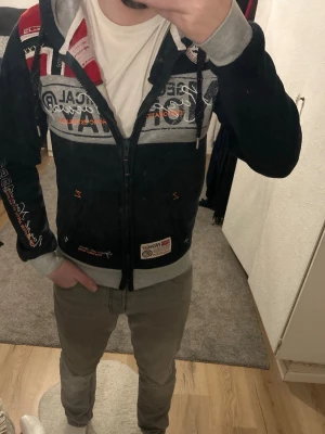 Geographical Norway hoodie med tryck - Mörkblå hoodie från Geographical Norway med dragkedja, gråa muddar och detaljer i rött, vitt och grått. Stor logga och patchar på bröstet och ärmen, samt norsk flagga på axeln. Mjuk insida och fickor framtill. Perfekt för dig som gillar streetwear och snygga detaljer.