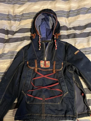 Mörkblå vindjacka från Napapijri 1/1 - Snygg mörkblå vindjacka från Napapijri med huva, halv dragkedja och dekorativa röda snören framtill. Jackan har norska flaggan som patch på bröstet, bruna läderdetaljer och broderad logga på ärmen. Perfekt för dig som vill ha en unik och sportig look.