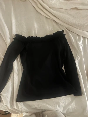 Svart off shoulder topp med volang - Trendig svart off shoulder topp med långa ärmar och volangdetalj längs urringningen. Toppens material är mjukt och stretchigt, vilket ger en snygg passform. Perfekt för dig som gillar en stilren men ändå lekfull look.