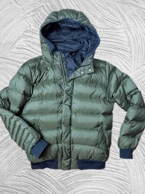 Mörkgrön ufferjacka från Scotch & Soda - Stilren grön pufferjacka med huva och mörkblå detaljer vid muddar och nederkant. Jackan har dragkedja och tryckknappar framtill samt två sidofickor. Perfekt för kalla dagar tack vare det vadderade materialet och den sportiga looken.