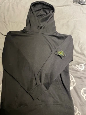 Svart hoodie från Stone Island - Säljer en svart hoodie från Stone Island med klassisk huva och känguruficka. På vänster ärm sitter det ikoniska Stone Island-märket i svart och gult. Hoodien är tillverkad i mjukt bomullsmaterial och har ribbade muddar vid ärmslut och nederkant. Perfekt för en clean och stilren look.