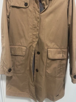 Beige trenchcoat från Levi's - Klassisk beige trenchcoat från Levi's med stora framfickor, knappar och rutigt innerfoder i blått och rött. Jackan har en rak passform, krage och detaljerade knappar vid ärmsluten. Perfekt för dig som gillar tidlös och stilren design.
