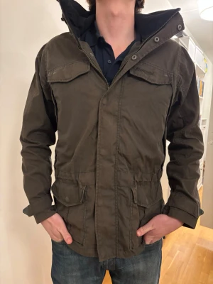 Grön vindjacka från Bergans - Snygg grön vindjacka från Bergans med fyra stora fickor framtill, justerbar huva och kardborre vid ärmsluten. Jackan har dragkedja och tryckknappar, samt diskret Bergans-logga på bröstet. Perfekt för blåsiga dagar och outdoor-äventyr.