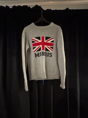 Grå stickad tröja  - Snygg grå stickad tröja från Morris med Union Jack-flagga och texten 'MORRIS' på bröstet. Tröjan har rund halsringning, ribbade muddar och nederkant. Perfekt för dig som gillar klassisk brittisk stil.