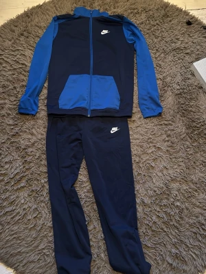 Nike blå träningsset med zip - Snyggt träningsset från Nike i mörkblått och klarblått. Jackan har hel dragkedja, ståkrage och två fickor framtill. Byxorna är matchande mörkblå med Nike-logga på låret. Perfekt för gymmet eller sportaktiviteter. Extremt sparsamt använd!🤗