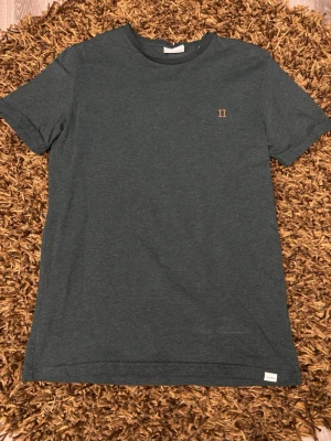 Mörkgrön t-shirt från Les Deux - Snygg mörkgrön t-shirt från Les Deux med diskret broderad logga i orange på bröstet. Klassisk rund halsringning och korta ärmar. Tillverkad i mjuk bomull för skön passform. Perfekt till jeans eller shorts.