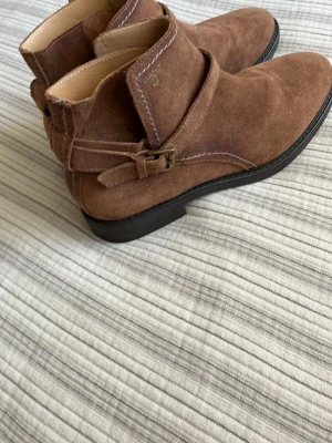 Bruna mockastövletter med spänne Gant - Snygga Gant bruna stövletter i mocka med dekorativt spänne på sidan och låg klack. Skorna har rundad tå och kontrastsömmar som ger en cool touch. Perfekta för dig som gillar stilrena boots med lite edge. Nyskick!