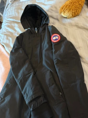 Svart parkas från Canada Goose - Säljer en svart parkasjacka från Canada Goose med klassisk logotyp på ärmen. Jackan har huva, dragkedja framtill och två stora fickor. Materialet är slitstarkt och vindtätt, perfekt för kalla dagar. Snygg och tidlös design som funkar till det mesta. och den har 2 stycken fickor inne🫶🏼