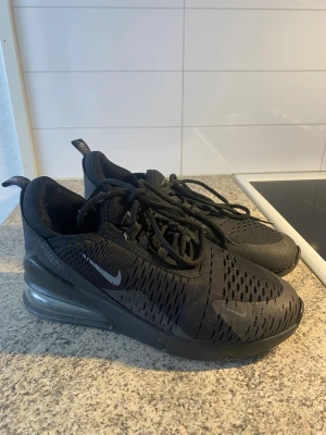 Nike Air Max 270 svarta sneakers - Säljer ett par helsvarta Nike Air Max 270 sneakers med mesh-överdel och synlig Air-enhet i hälen. Skorna har en sportig look med små vita Nike-loggor på sidan och tungan. Perfekta för dig som gillar stilrena och bekväma sneakers. Står storlek 38 men passar flera. 