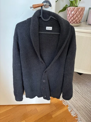 Mörkgrå kofta från John Henric - Snygg mörkgrå kofta från John Henric med bred sjalkrage och knäppning framtill. Koftan är ribbstickad och har långa ärmar, perfekt för lager på lager. Klassisk och stilren design som passar till det mesta.