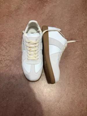 Maison margiela gats - Snygga vita sneakers från Maison Margiela med beige mockadetaljer och klassisk gummisula. Skorna har en retroinspirerad design med rund tå, vita skosnören och diskreta paneler i både läder och mocka. Perfekta för dig som gillar stilrena och tidlösa sneakers.