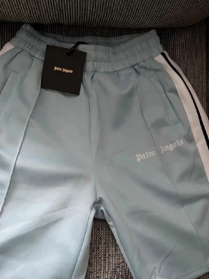 Ljusblå shorts från Palm Angels - Säljer ett par ljusblå shorts från Palm Angels med vita och svarta ränder längs sidorna. Shortsen har elastisk midja, sidofickor och logga broderad på vänster ben. Perfekta för en avslappnad och sportig stil.