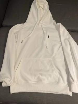 Vit hoodie från Polo Ralph Lauren - Snygg vit hoodie från Polo Ralph Lauren med klassisk huva och dragsko. Har en känguruficka framtill och den ikoniska lilla broderade loggan på bröstet. Tillverkad i mjukt material som känns skönt mot huden. Perfekt för en clean och stilren look.