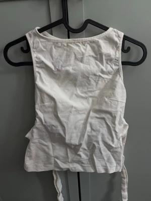 Vit croppad linnetopp med knytband - Säljer en vit croppad linnetopp med breda axelband och knytband i ryggen. Toppen har en enkel, stilren design och är tillverkad i mjukt material. Perfekt för att styla med höga jeans eller kjol.