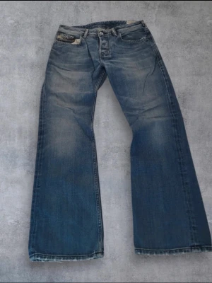  diesel bootcut jeans - Säljer ett par blåa bootcut jeans med snygga slitningar och slitna benslut. Jeansen har klassiska fem fickor och en cool tvätt med ljusare partier framtill. Perfekta för dig som gillar en avslappnad och trendig look.