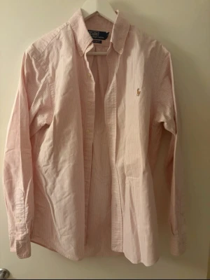 Rosa och vit randig skjorta Polo Ralph Lauren - Klassisk rosa och vit randig skjorta från Polo Ralph Lauren med broderad logga på bröstet. Skjortan har lång ärm, knappar framtill och är gjord i mjuk bomull. Perfekt för en clean och stilren look.