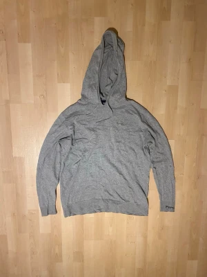 Grå hoodie från Pepe Jeans - Snygg grå hoodie från Pepe Jeans med broderad logga på bröstet och diskret text på ärmen. Klassisk passform med stor huva och ribbade muddar. Tillverkad i mjukt bomullsmaterial som känns skönt mot huden.