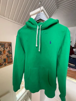 Grön hoodie från Polo Ralph Lauren - Snygg grön hoodie från Polo Ralph Lauren med klassisk känguruficka och vit dragsko i huvan. Ikoniska loggan på bröstet. Tillverkad i mjuk bomull som känns skön mot huden. Perfekt för en chill och avslappnad stil. Defekter på bild 4-5.                                                            Byten kan vara intressant!