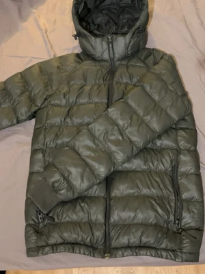 Mörkgrön dunjacka Peak Performance - Snygg mörkgrön dunjacka från Peak Performance med huva och dragkedja framtill. Jackan har två sidofickor med dragkedja och diskret logga på bröstet samt ärmen. Perfekt för kalla dagar och riktigt skön att bära. Använd ett par gånger bara.