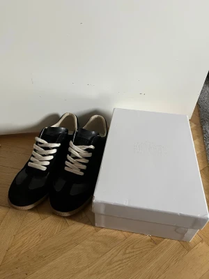 Svarta Maison Margiela Gats - Svarta sneakers från Maison Margiela med vita snören. Skorna har en klassisk låg siluett och gummisula med mönster undertill. Materialet är en mix av mocka och läder för en clean och stilren look. Dustbag och box finns. Skorna är använda en gång så de är nästan som nya. Priset ej hugget i sten vid snabb affär