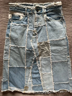 Patchwork jeanskjol med fransar - Unik patchwork jeanskjol i ljusblå denim med råa fransade kanter och synliga sömmar. Kjolen har klassiska fickor fram och bälteshällor i midjan. Perfekt för dig som gillar en edgy och kreativ stil.