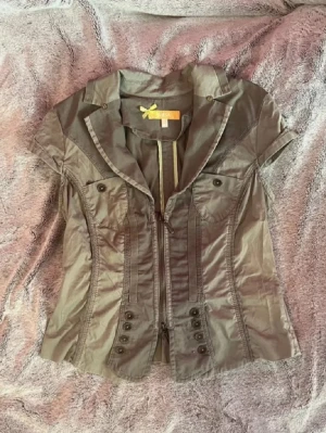 Tröja - Figursydd modell med dekorativa knappar framtill, fickor med lock och krage. Dragkedja framtill och volangdetalj baktill ger en cool och unik look. .