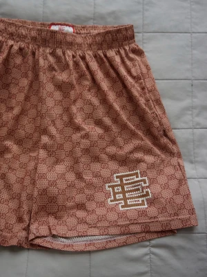 Eric Emmanuel | Shorts - Säljer ett par beige shorts från GG x Eric Emmanuel NYC med all-over GG-mönster och en vit broderad detalj på ena benet. Shortsen har elastisk midja och är tillverkade i ett lätt material som passar perfekt till sommaren. OBS: storlek L men passar M 