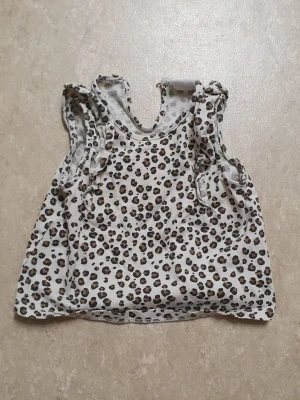 Topp - Säljer en söt ärmlös topp med leopardmönster i svart och brunt på vit botten. Toppen har volangdetaljer vid axlarna och en rund halsringning. Perfekt för dig som vill sticka ut lite extra med ett trendigt djurmönster.