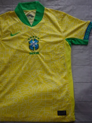 Brasil | Nike fotbollströja - Gul Brasilien fotbollströja från Nike med gröna detaljer och CBF-emblem på bröstet. Tröjan har ett grafiskt mönster över hela tyget, grönt Nike-märke och grön krage. Perfekt för dig som älskar fotboll och vill stötta Brasilien. Helt ny och oanvänd! 