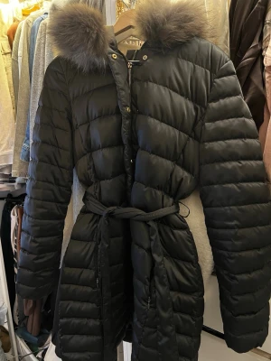 Hugo boss jacka med äkta päls - Snygg svart lång vinterjacka med bälte i midjan och stor huva med fluffig äkta päls. Jackan har quiltad design och stängs med dragkedja och knappar. Perfekt för kalla vinterdagar och ger en trendig siluett.