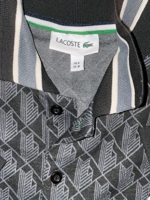 Mönstrad grå skjorta från Lacoste - Snygg grå skjorta från Lacoste med grafiskt mönster i vitt och svart. Skjortan har klassisk krage med vita och grå ränder, knappar framtill och en bröstficka. Perfekt för dig som gillar stilrena detaljer och ikonisk design.