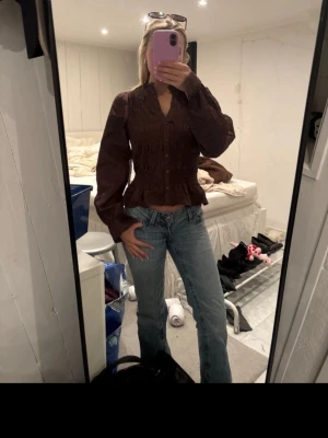 Brun blus med smock och volang - Snygg brun blus från bubbleroom  med smockdetaljer över bröstet och volangkant nedtill. Blusen har v-ringning, knappar framtill och långa puffiga ärmar med resår vid handlederna. Perfekt att styla med jeans eller kjol för en trendig look.