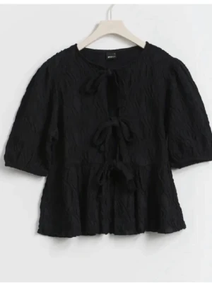 Svart blus med knyt och struktur - Trendig svart blus med tre dekorativa knyt framtill och lätt puffade ärmar. Toppen har en strukturerad yta och avslappnad passform som ger en cool och modern look. Perfekt att styla med både kjol och jeans.