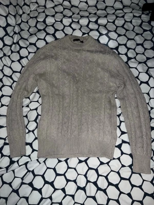 Beige kabelstickad tröja - Säljer en beige kabelstickad tröja med rund hals och ribbade muddar vid ärmslut och nederkant. Tröjan har ett klassiskt flätmönster och är perfekt för dig som gillar en stilren och mysig look. Passar till både jeans och kjol.