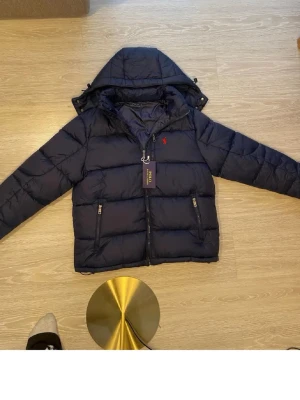Mörkblå pufferjacka från Polo Ralph Lauren - Snygg mörkblå pufferjacka från Polo Ralph Lauren med avtagbar huva och två dragkedjeförsedda fickor framtill. Jackan har det klassiska röda Polo-logot broderat på bröstet och en quiltad design som håller värmen. Perfekt för kalla vinterdagar. Jackan är ej äkta men det är inte några Ralph Lauren jackor på Plick.