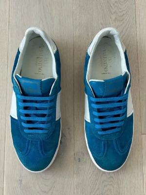 Valentino flycrews  - Snygga blåa sneakers från Valentino med vita detaljer och blå mocka. Skorna har vita partier i läder, blå skosnören och coola nitar på hälen. Klassisk låg modell som passar till många olika stilar. Tillverkade i Italien. Endast 1 nit som är av… syns inte vid användning, storlek 42 passar även 43. Skriv vid fundering!