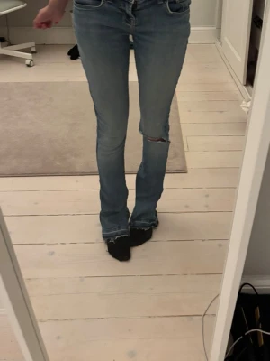 Ljusblå bootcut jeans med slitning - Säljer ett par ljusblå bootcut jeans med slitning över knät och råa kanter vid benslut. Jeansen har klassisk femficksmodell och normal midja. Perfekta för en avslappnad och trendig look.