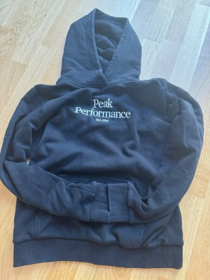 Svart hoodie från Peak Performance - Snygg svart hoodie från Peak Performance med broderad logga och text 'Est. 1986' på bröstet. Klassisk modell med stor huva och magficka. Perfekt för chill dagar eller när du vill ha en clean streetwear-look. Går för 1000kr ny