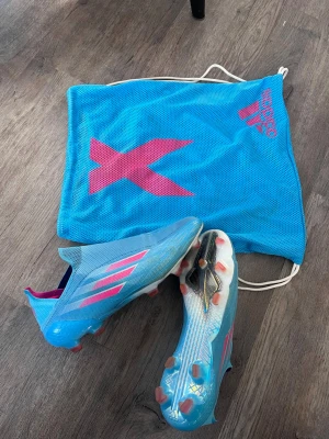 Blå Adidas X Speedflow fotbollsskor - Säljer ett par blå Adidas X Speedflow fotbollsskor med rosa detaljer och vita sulor. Skorna har en strömlinjeformad design utan snörning och är tillverkade i lätt syntetmaterial för maximal fart på planen. Medföljer matchande blå påse med rosa X-logga.