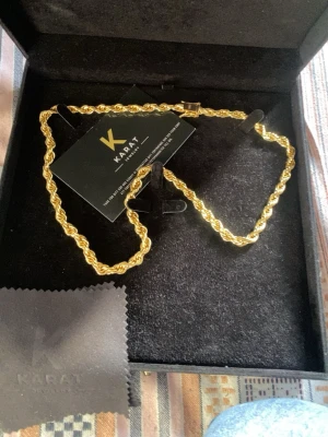 Guldfärgat repkedje-halsband Karat - Snyggt guldfärgat halsband från Karat med repkedje-design. Halsbandet har en robust låsdetalj med Karat-logga och levereras i en elegant svart ask. Perfekt accessoar för att lyfta din stil med en lyxig touch.