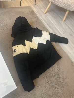 Svart dunjacka från Moose Knuckles - Säljer en svart dunjacka från Moose Knuckles med vita detaljer och ikonisk logga på ärmen. Jackan har huva, dragkedja framtill och två fickor med dragkedja. Tjock och varm design, perfekt för kalla vinterdagar.