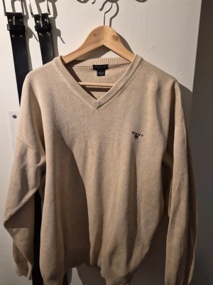 Beige stickad tröja från Gant - Klassisk beige stickad tröja från Gant i 100% bomull. Tröjan har V-ringning, ribbade muddar och diskret Gant-logga broderad på bröstet. Perfekt för en stilren och avslappnad look.